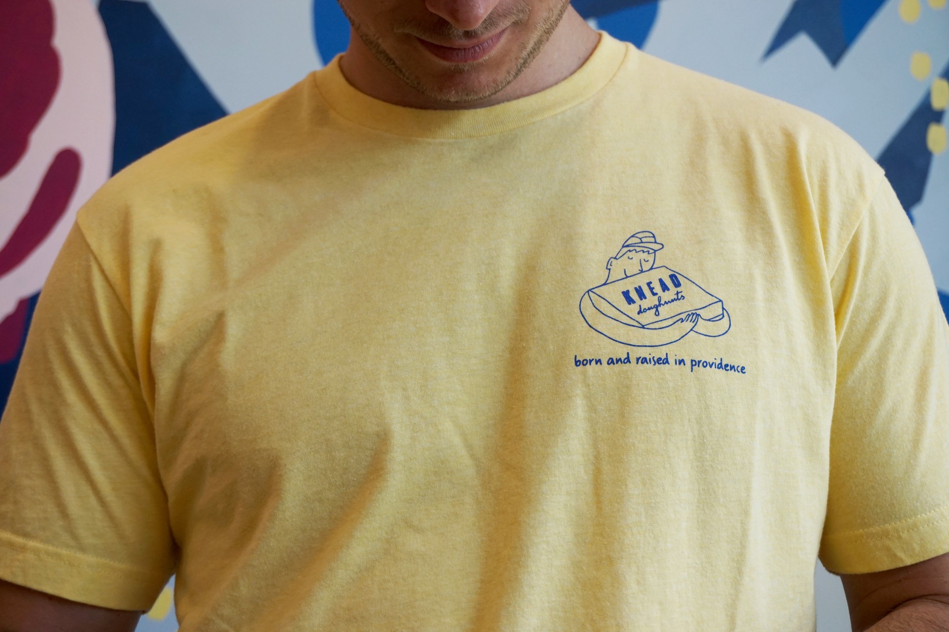 Model wearing yellow short sleeve shirt. Pocket design is of character hugging a box of doughnuts with text underneath that reads, "Made in Providence" 