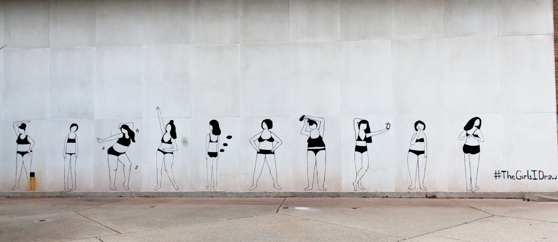 Photograph of wall with mural. Mural consists of 10 women in undergarments in various poses. Women are all rendered in black lines made out of duct tape. 