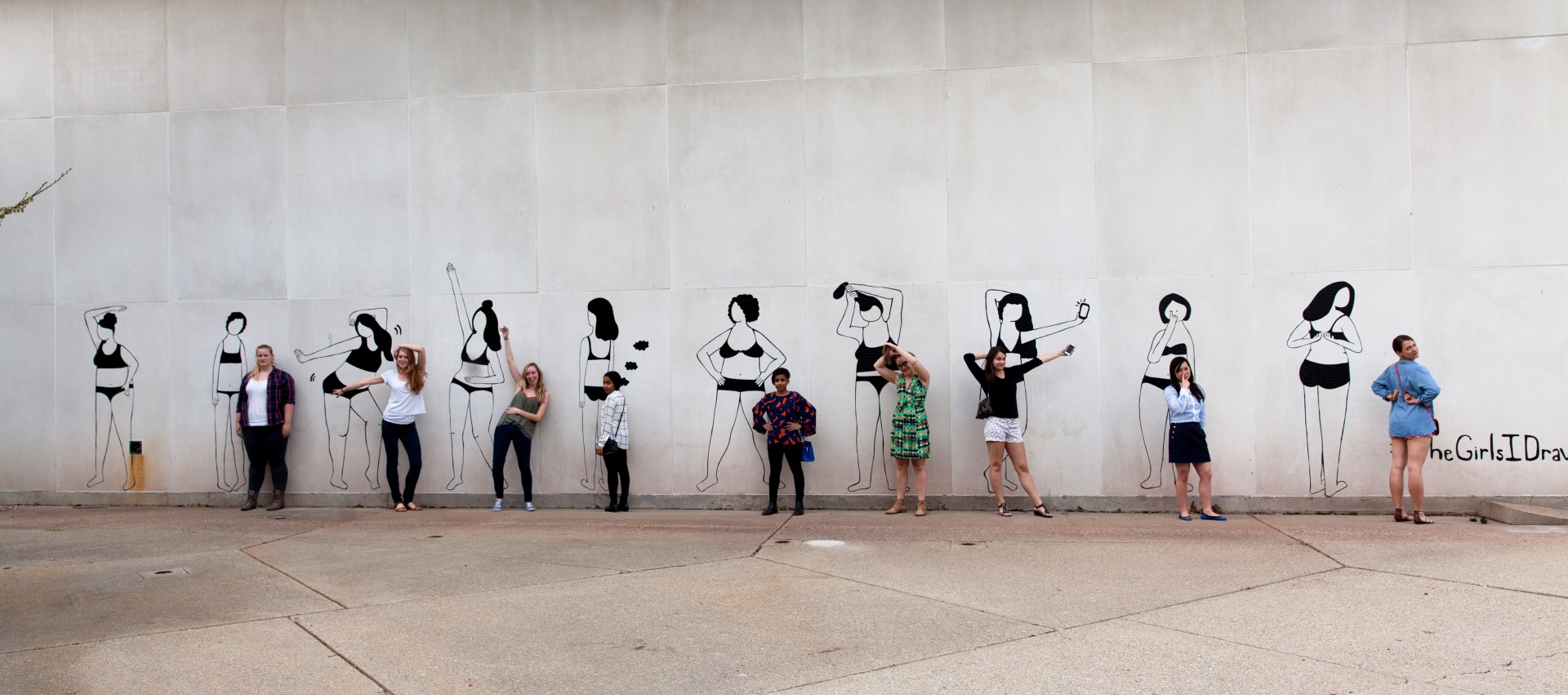 Photograph of wall with mural. Mural consists of 10 women in undergarments in various poses. Women are all rendered in black lines made out of duct tape. People are posed in front of each figure in mural mimicking the pose. 