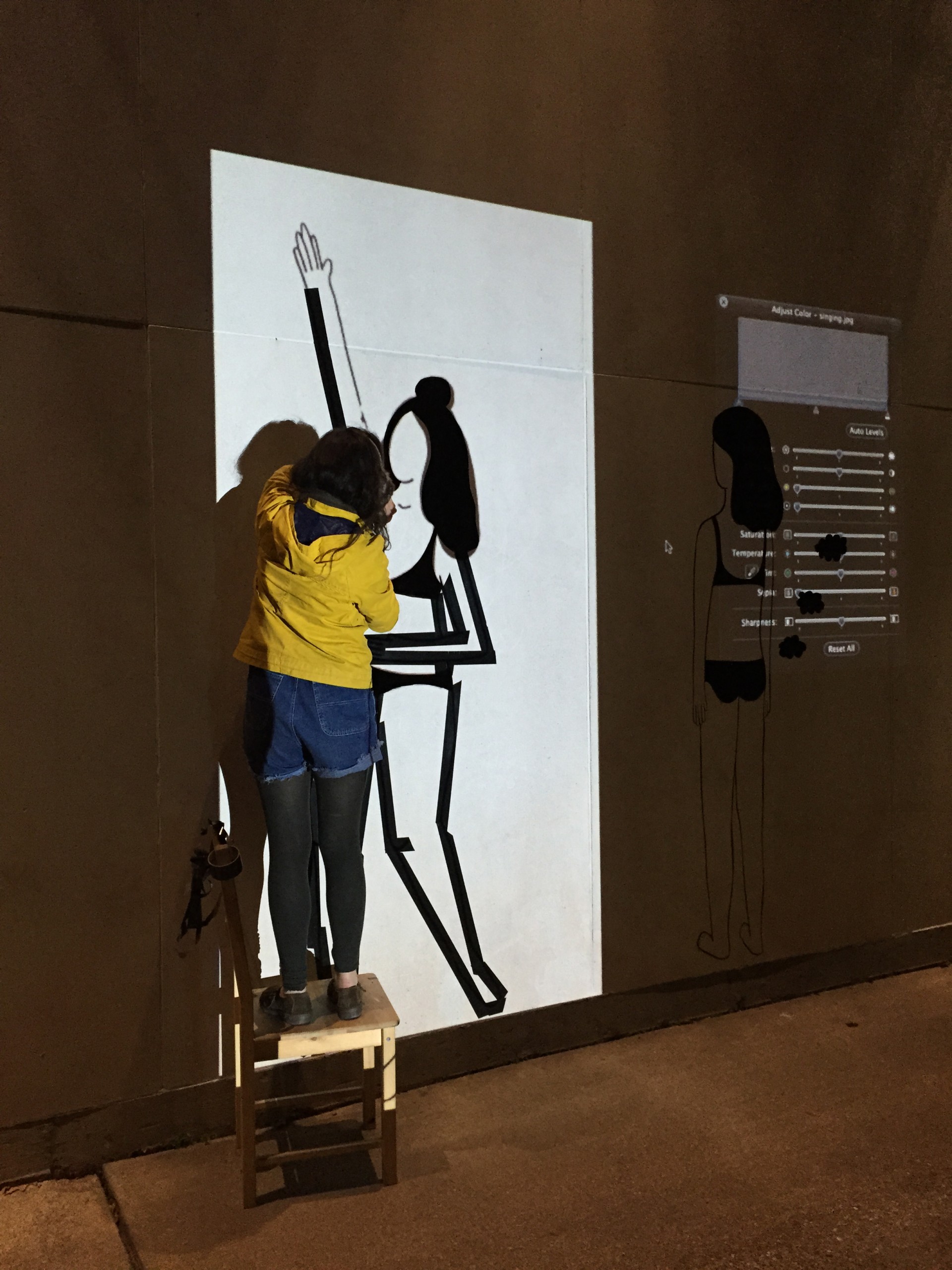 Image of artist making duct tape mural at night. She is standing on chair cast in light from projected image onto wall. 