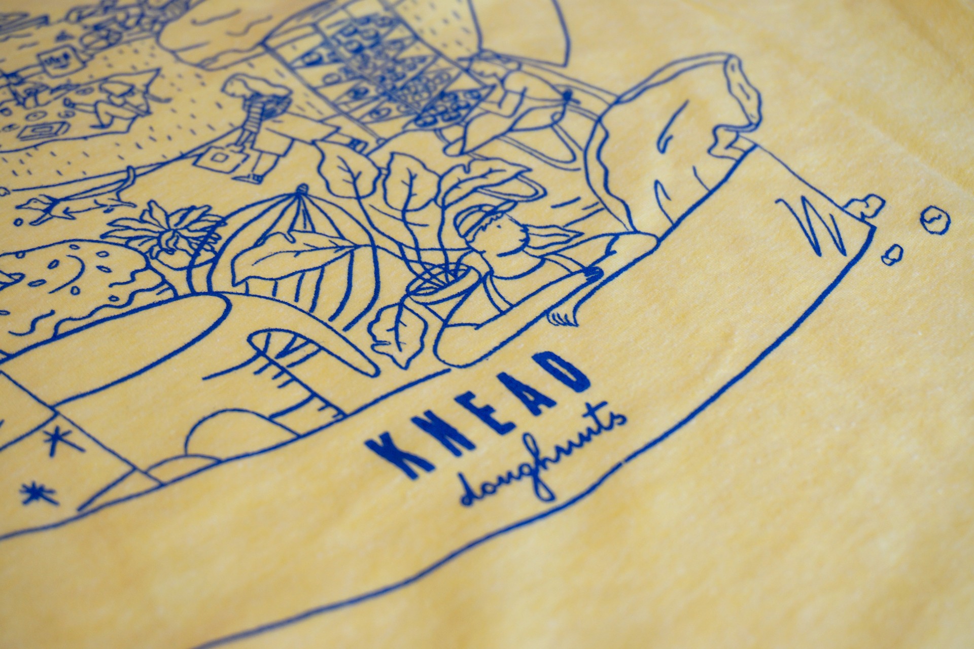 Detail of full back design of yellow t shirt. Text reads "Knead Doughnuts" Illustration shows oversized baking items, Knead employees and plants all inside Knead box of doughnuts. 