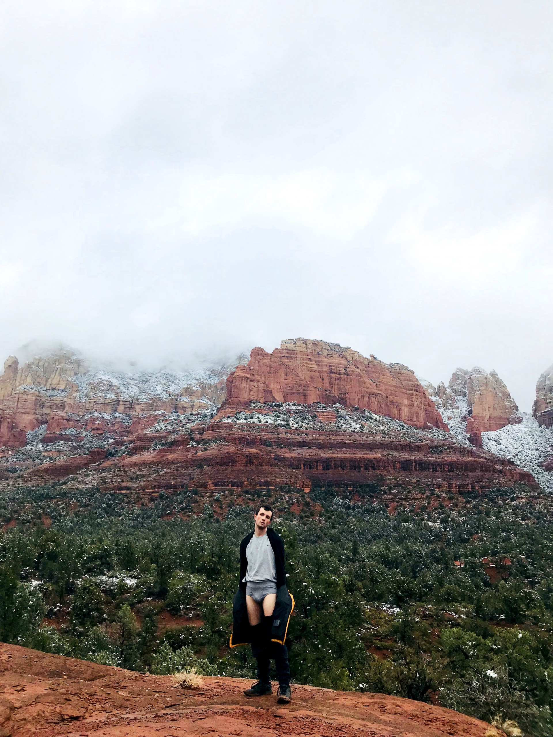 “Photograph of snow topped canyon in Sedona Arizona. Landscape covered in mist. Male model stands in center foreground of image with pants pulled below waist exposing grey underwear.”