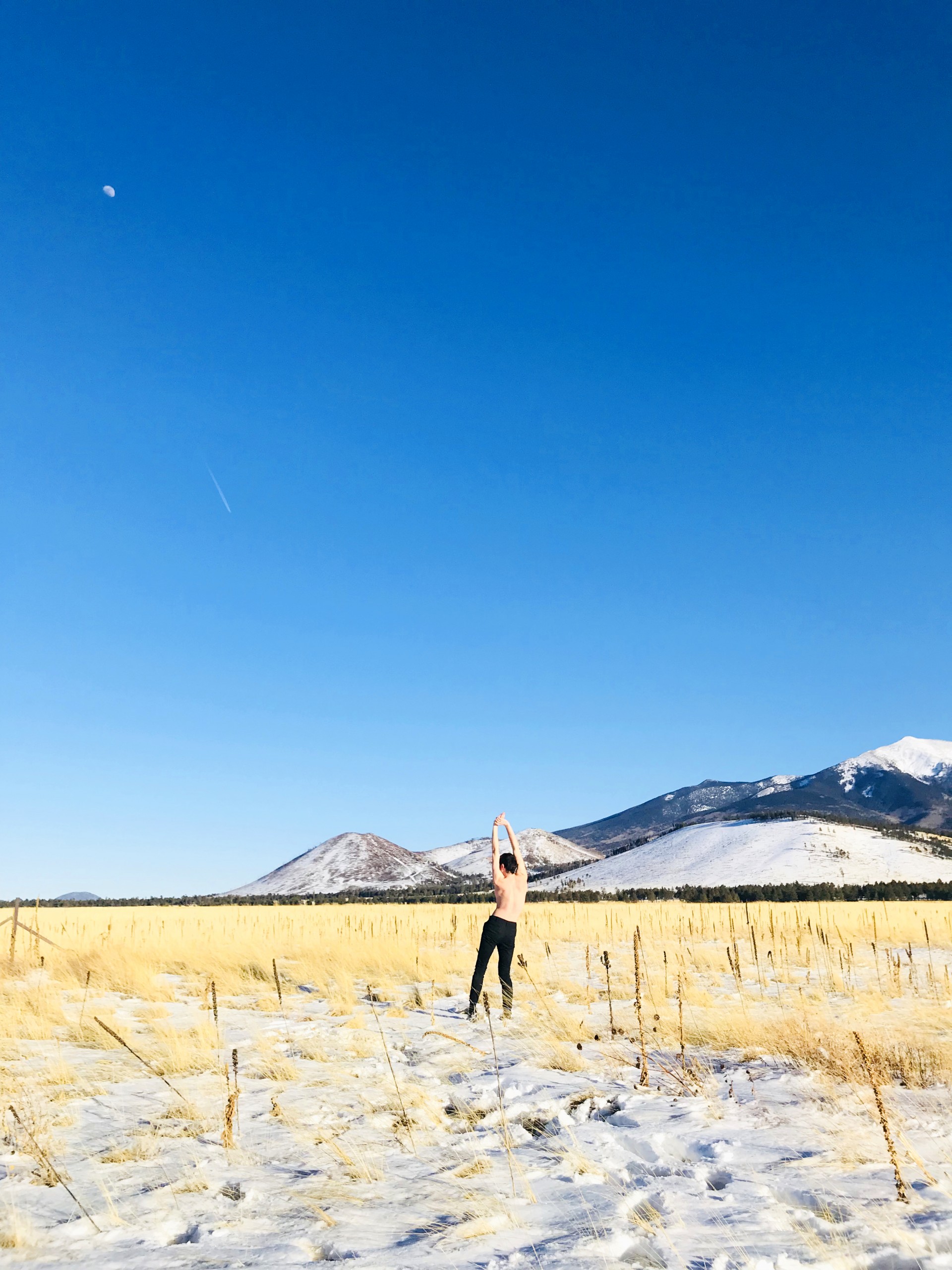 Photograph of snowy wheat field. Male model stands with back towards viewer in center of image. Body is slightly curved. Sky is blue. 