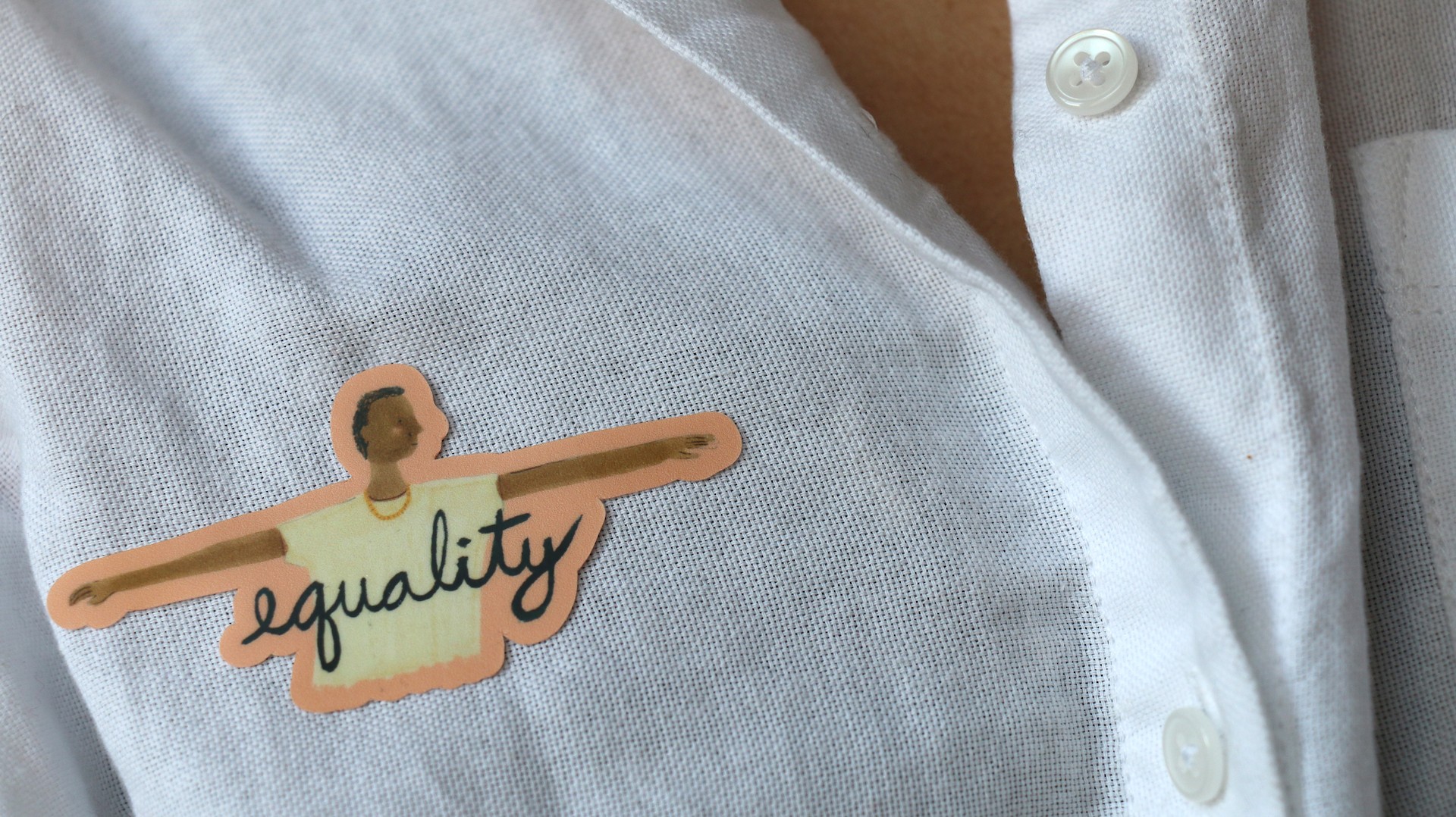 Sticker placed on white shirt. Sticker is of young man with arms outstretched to either side with text that reads "equality"
