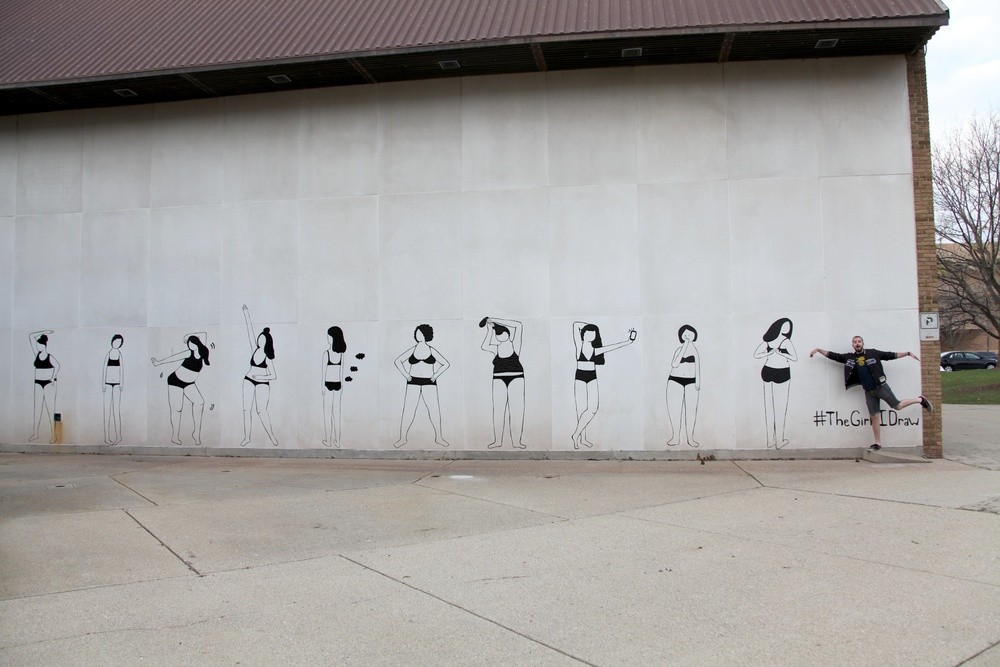 Photograph of wall with mural. Mural consists of 10 women in undergarments in various poses. Women are all rendered in black lines made out of duct tape. Single person poses to right side of mural. 
