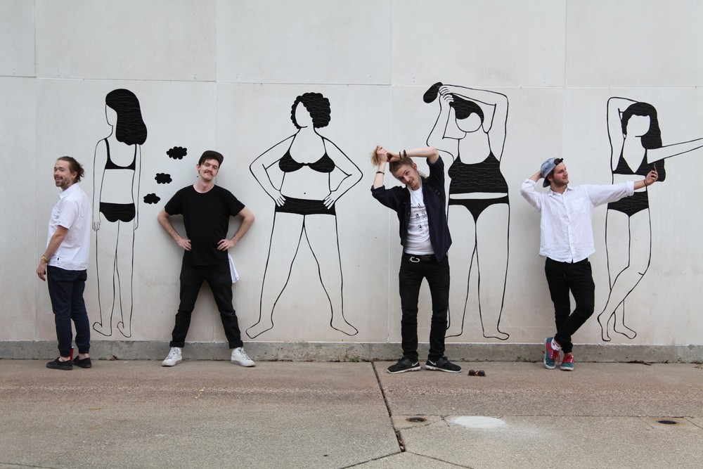 Photograph of wall with mural. Photograph shows 4 women in undergarments in various poses. Women are all rendered in black lines. People are posed in front of each figure in mural mimicking the pose. 