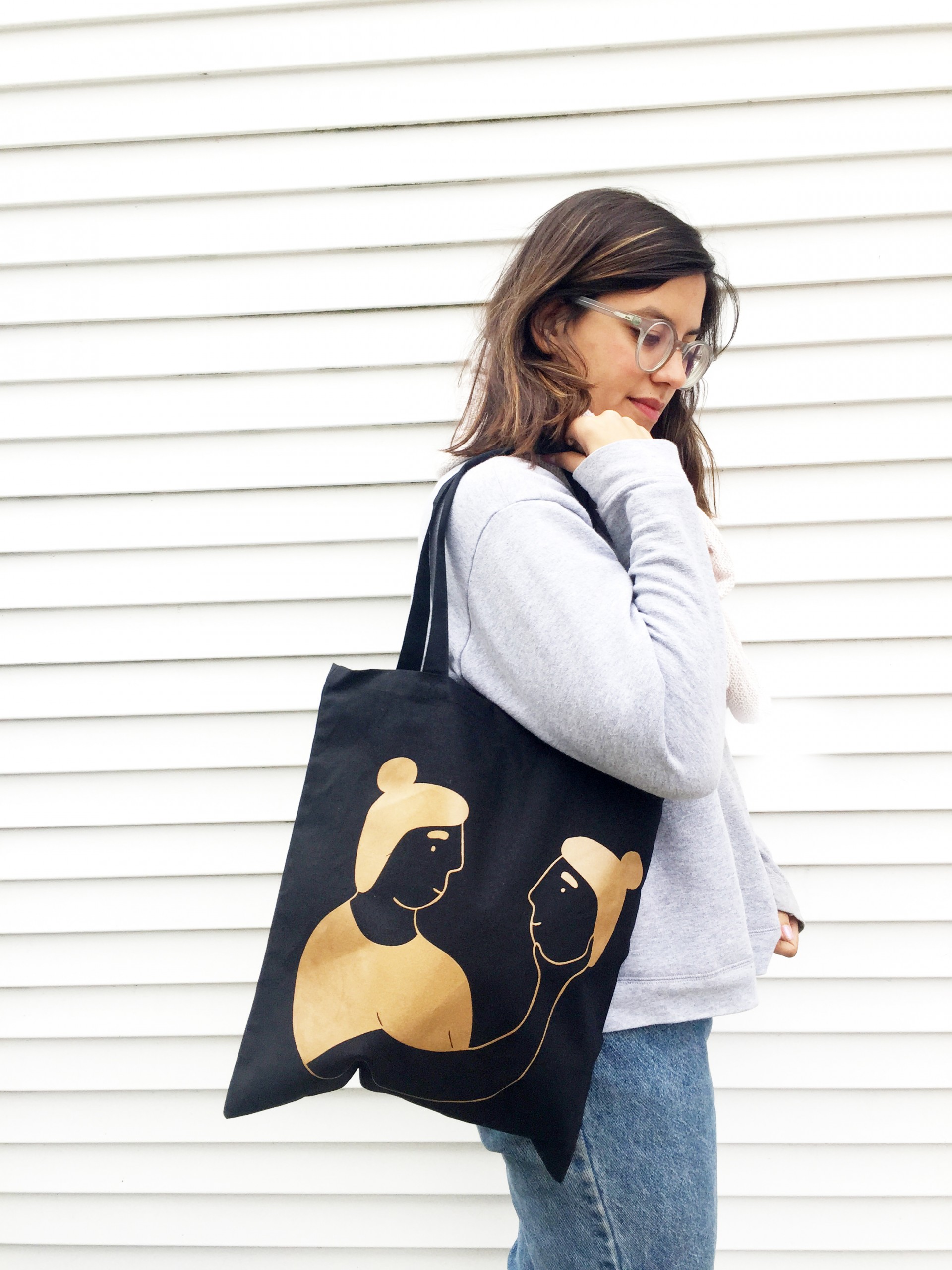 Black tote bag with girl printed in gold who is smiling at a head she is holding in her hand 